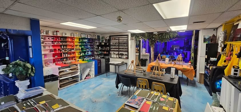The A.I.M. art studio: shelves of paint arranged by color along one wall, easels set up on tables, student artwork laid out in the foreground and a lit studio space beyond.