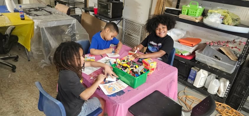 Three young Lion Hearts members sitting around a table in the studio, coloring and working with a tray of craft materials between them.