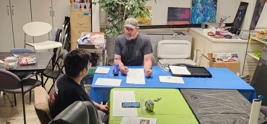 Instructor Alan Voyles sitting at a table with a student, going over a hand-drawn layout on paper with a tape measure and ruler beside it.