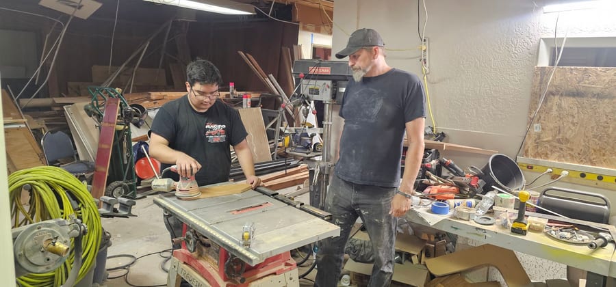 A student sanding a board at a table saw in the A.I.M. shop while an instructor watches.